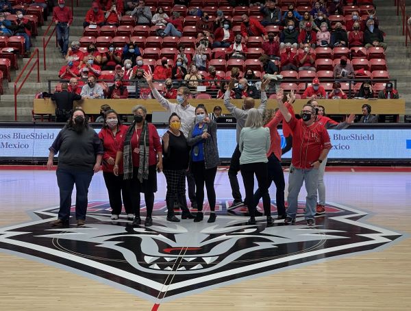 A group of SHAC staff pose on the floor of the UNM Arena and wave to the crowd of spectators at the game.
