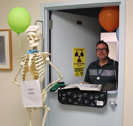 An X-Ray technician stands in the doorway of SHAC Radiology. A skeleton with a bow tie is nearby.