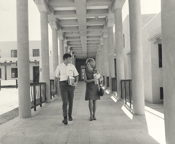 A male and female student are walking under the SHAC portal. Each carries books.